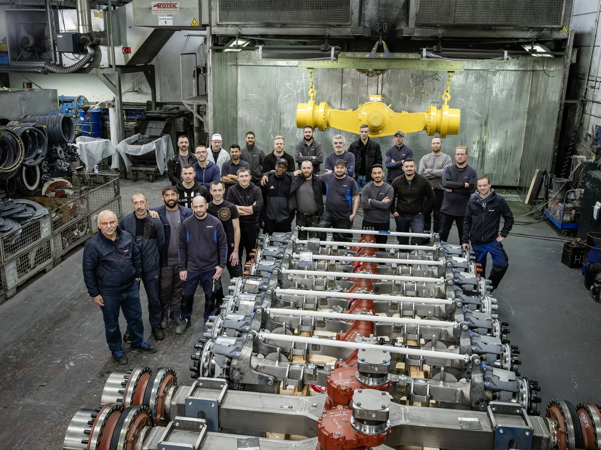 Group of workers in a manufacturing or industrial facility standing around machinery parts, with a large yellow mechanical component suspended overhead.