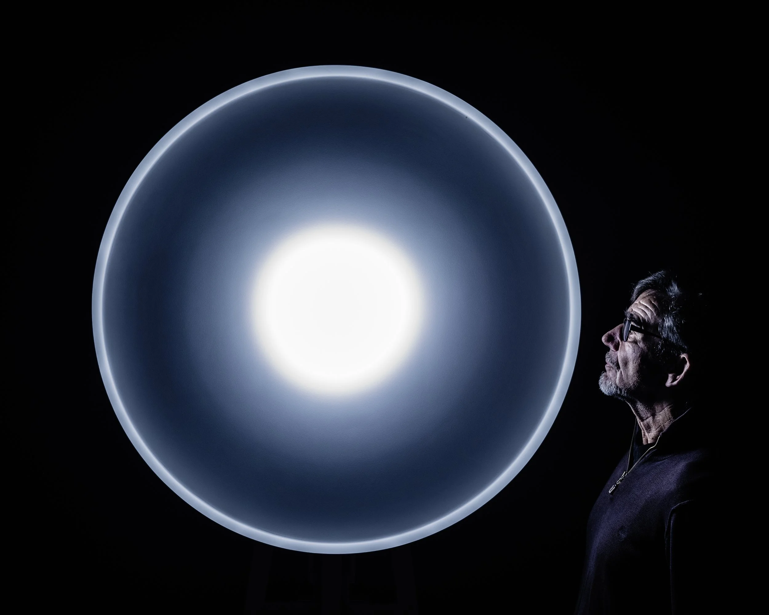 A side profile of a man with glasses and gray hair looking at an illuminated circular object in a dark environment.