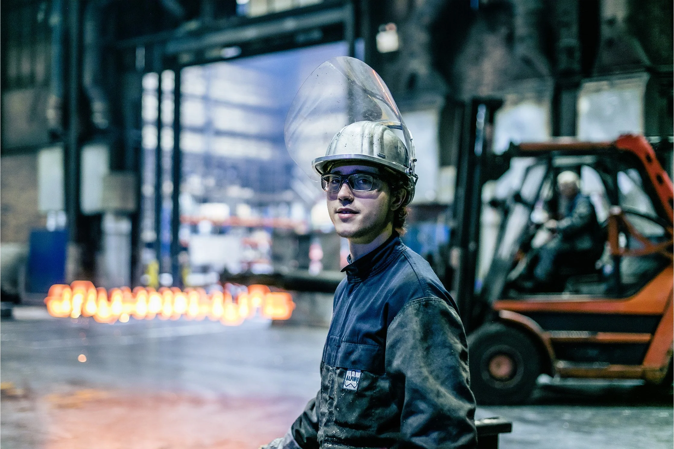 A young man wearing glasses and a firefighter helmet stands inside a garage or workshop filled with tools and equipment. The background shows a blurred sign with the hashtag '#CHAMPAGNE' and blurred machinery.