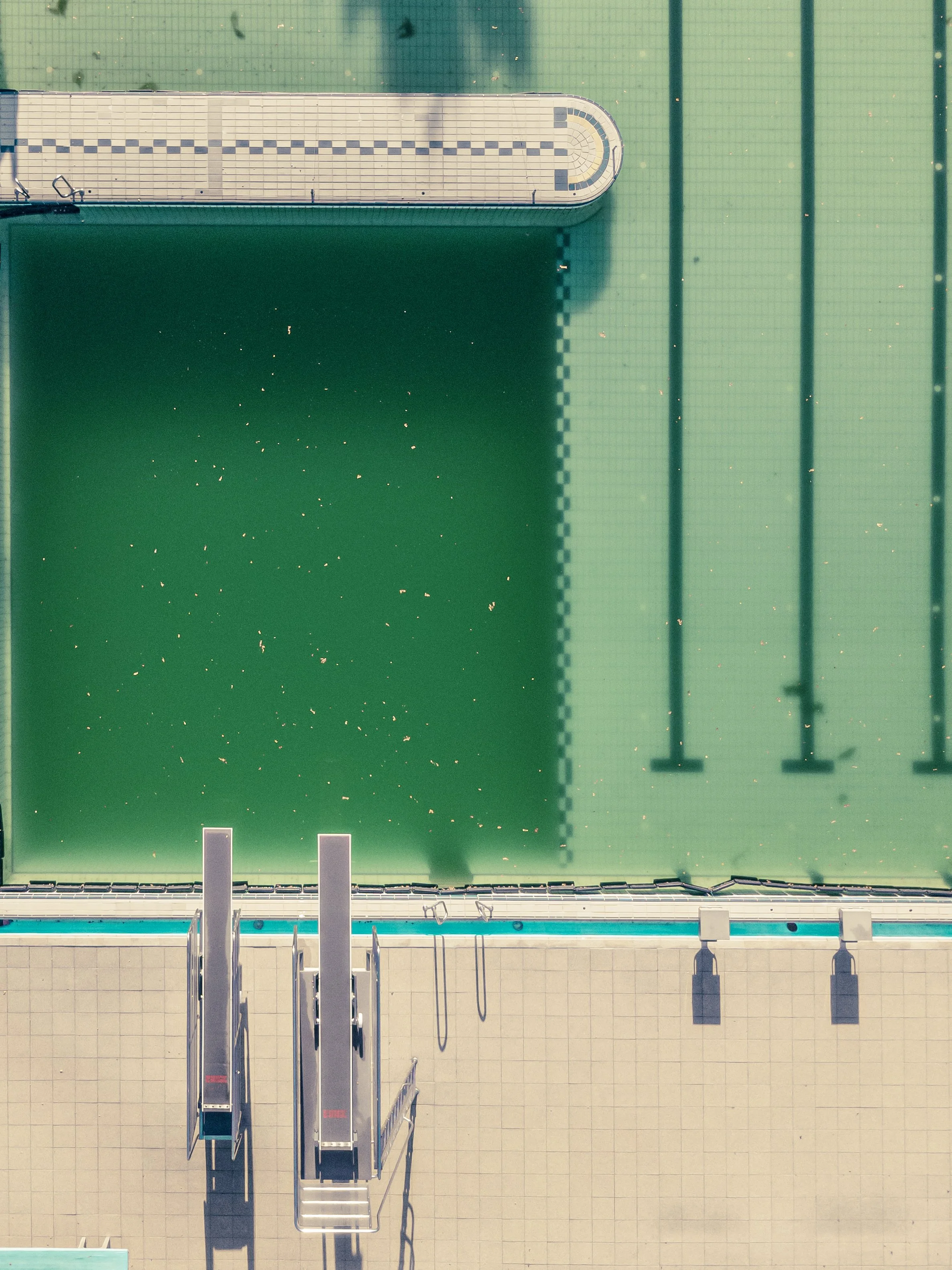 An aerial view of a swimming pool with multiple lanes and diving blocks, surrounded by a beige-tiled deck.