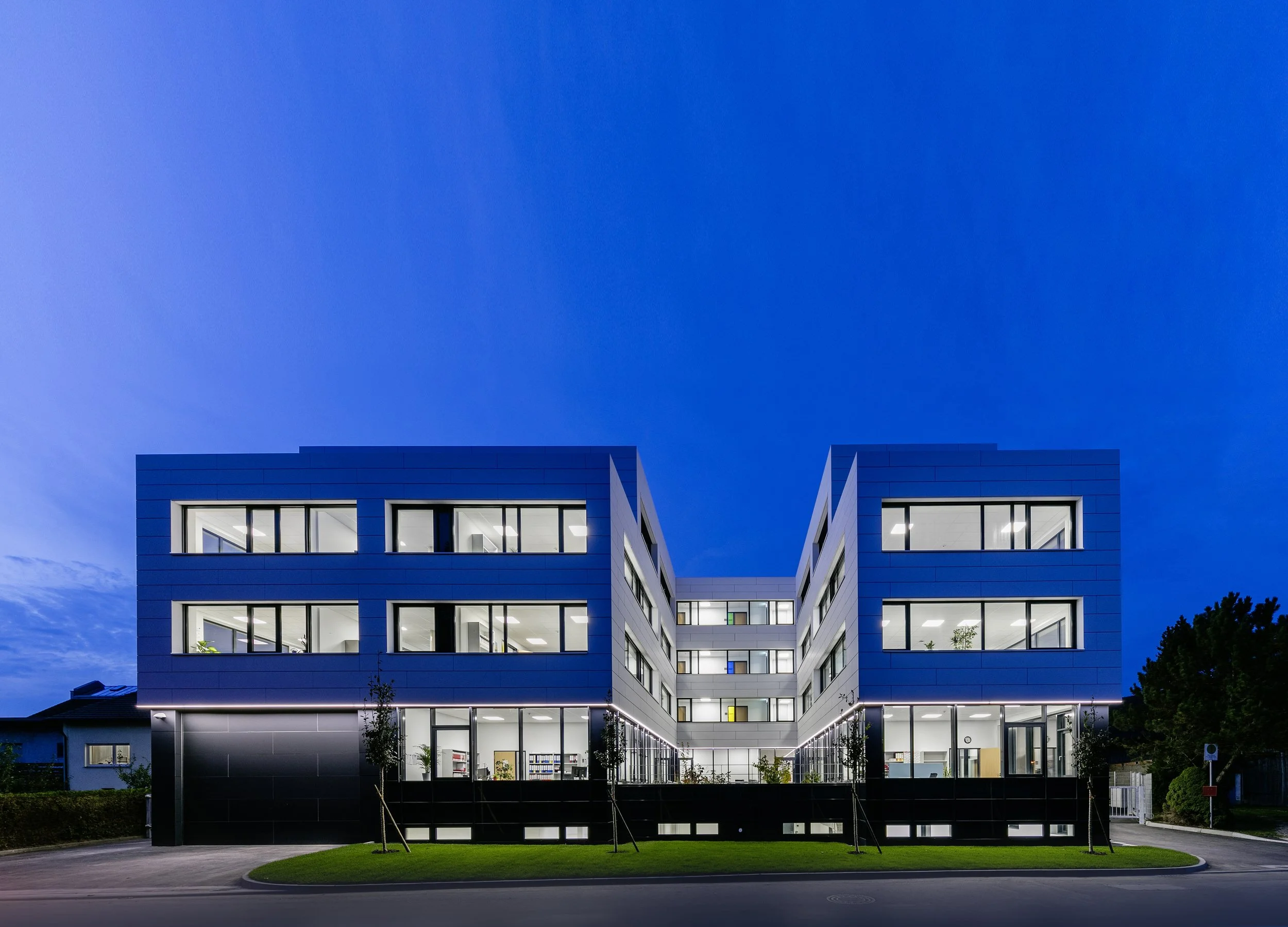 Modern multi-story office building illuminated at night with blue exterior walls and large glass windows, situated on a curved road with trees and grass.