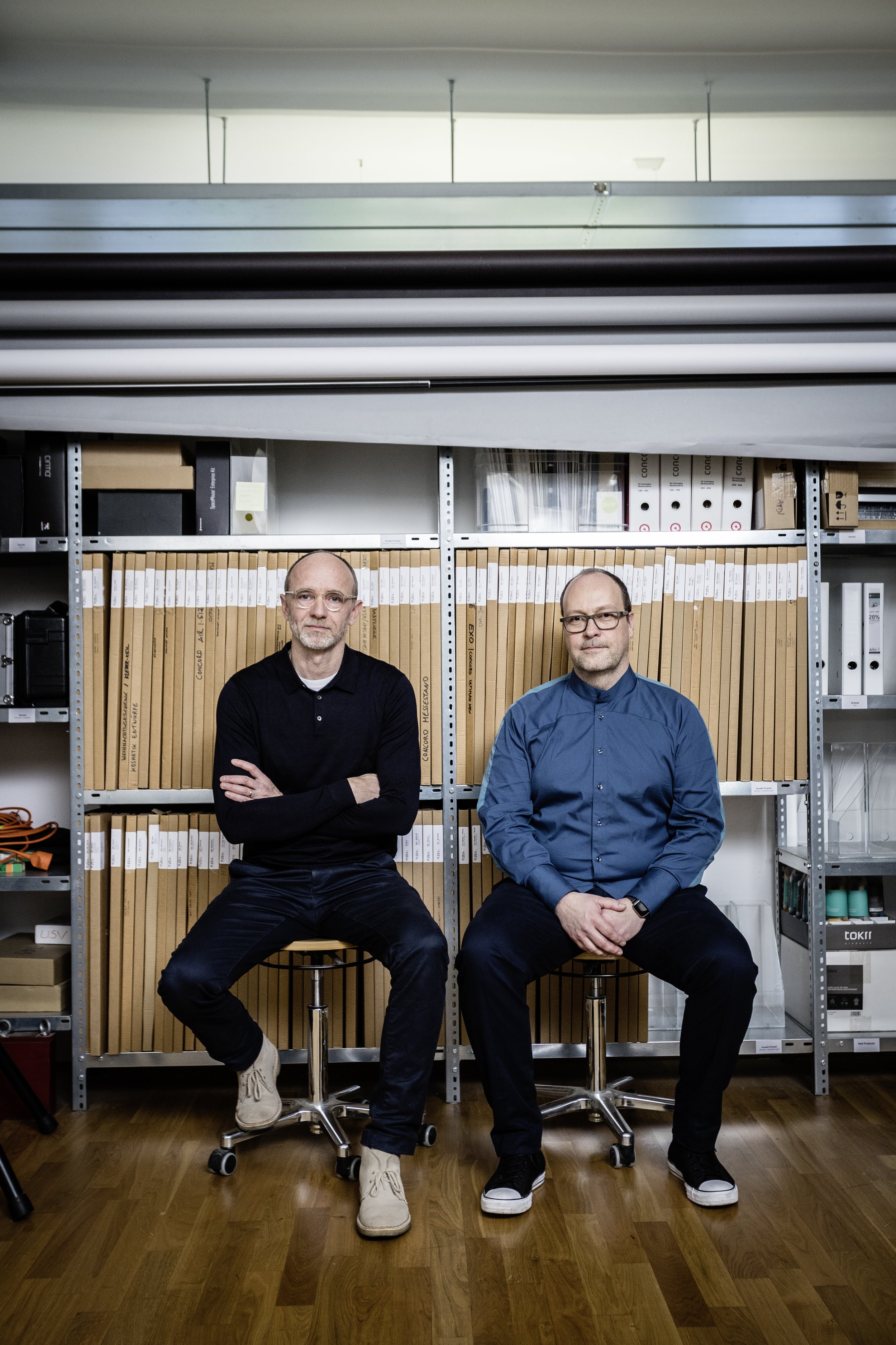 Two men sitting on stools in front of a metal shelving unit filled with binders and office supplies, in a room with hardwood floor.