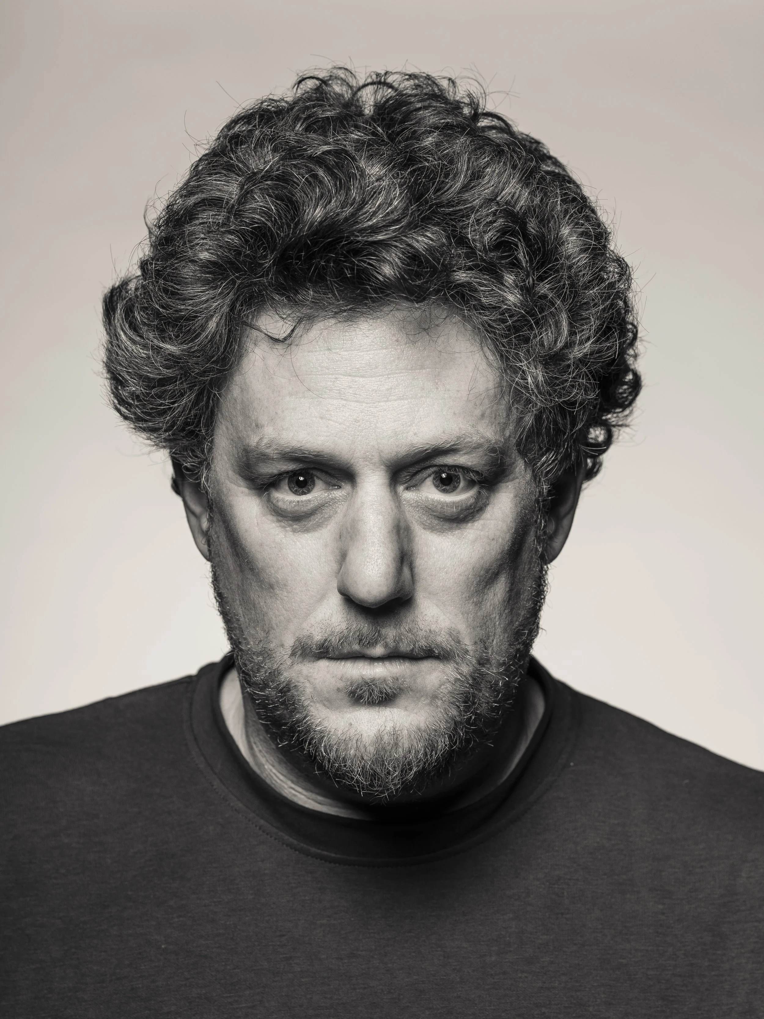 Close-up black-and-white portrait of a man with curly hair and a beard, wearing a dark t-shirt, against a plain background.