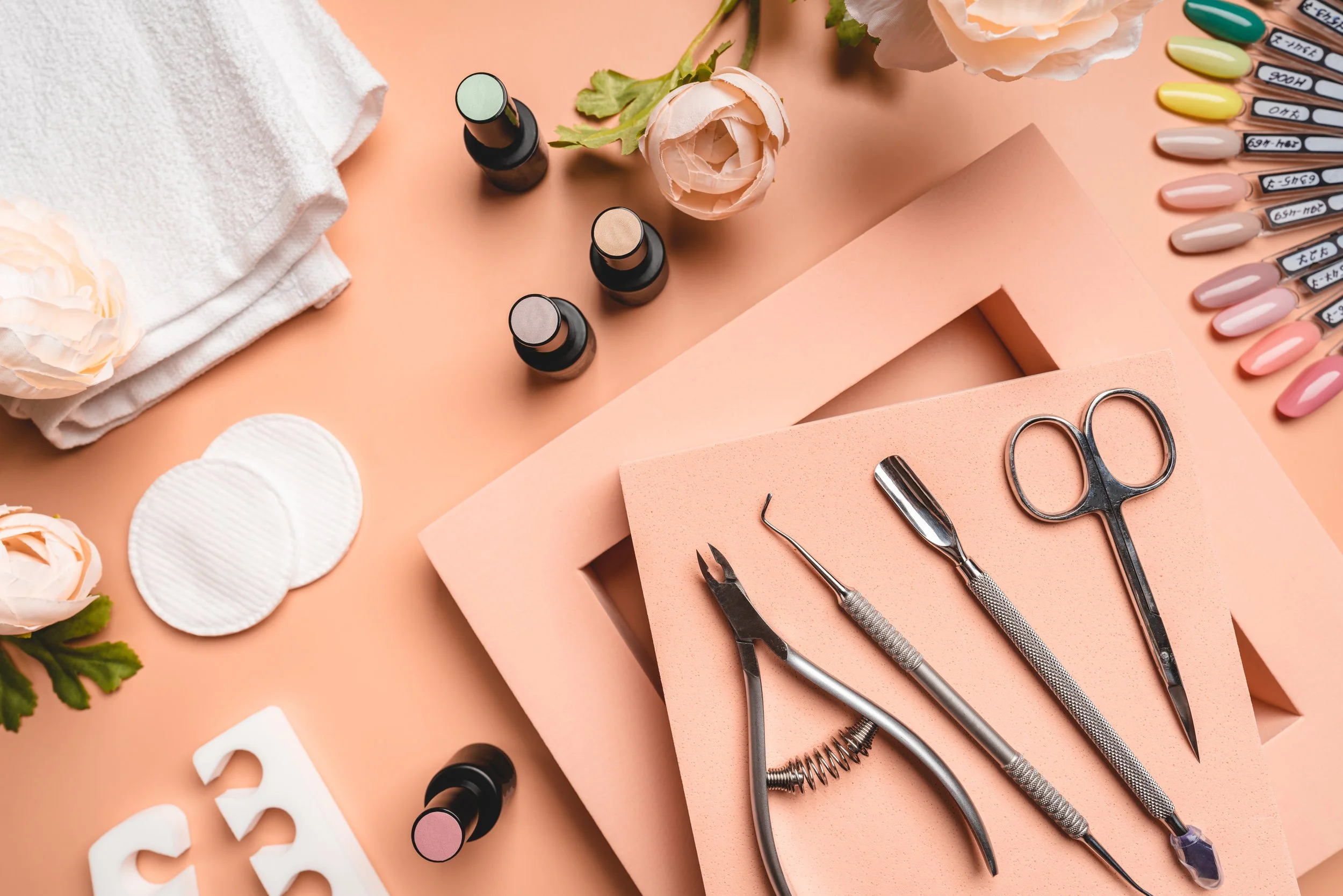 Manicure tools including cuticle nippers, an orange stick, scissors, and tweezers on pink foam, with nail polish bottles, nail swatches, flowers, and cotton pads on a peach-colored background.