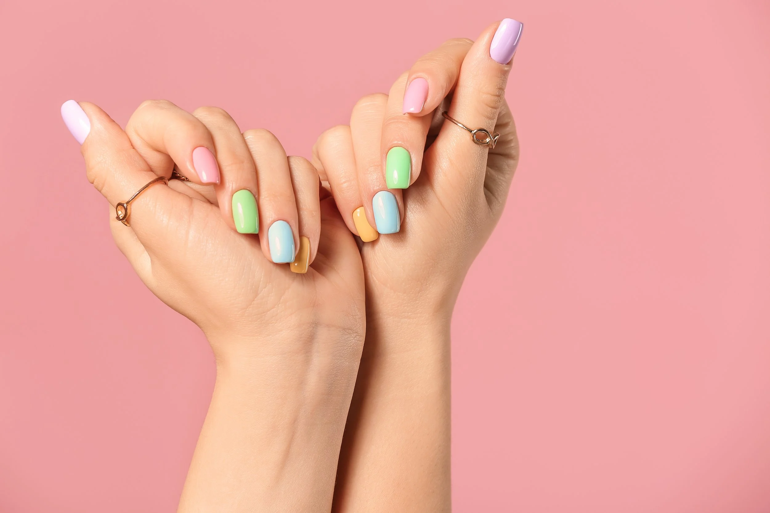 Close-up of a woman's hands with pastel-colored nail polish, set against a pink background.