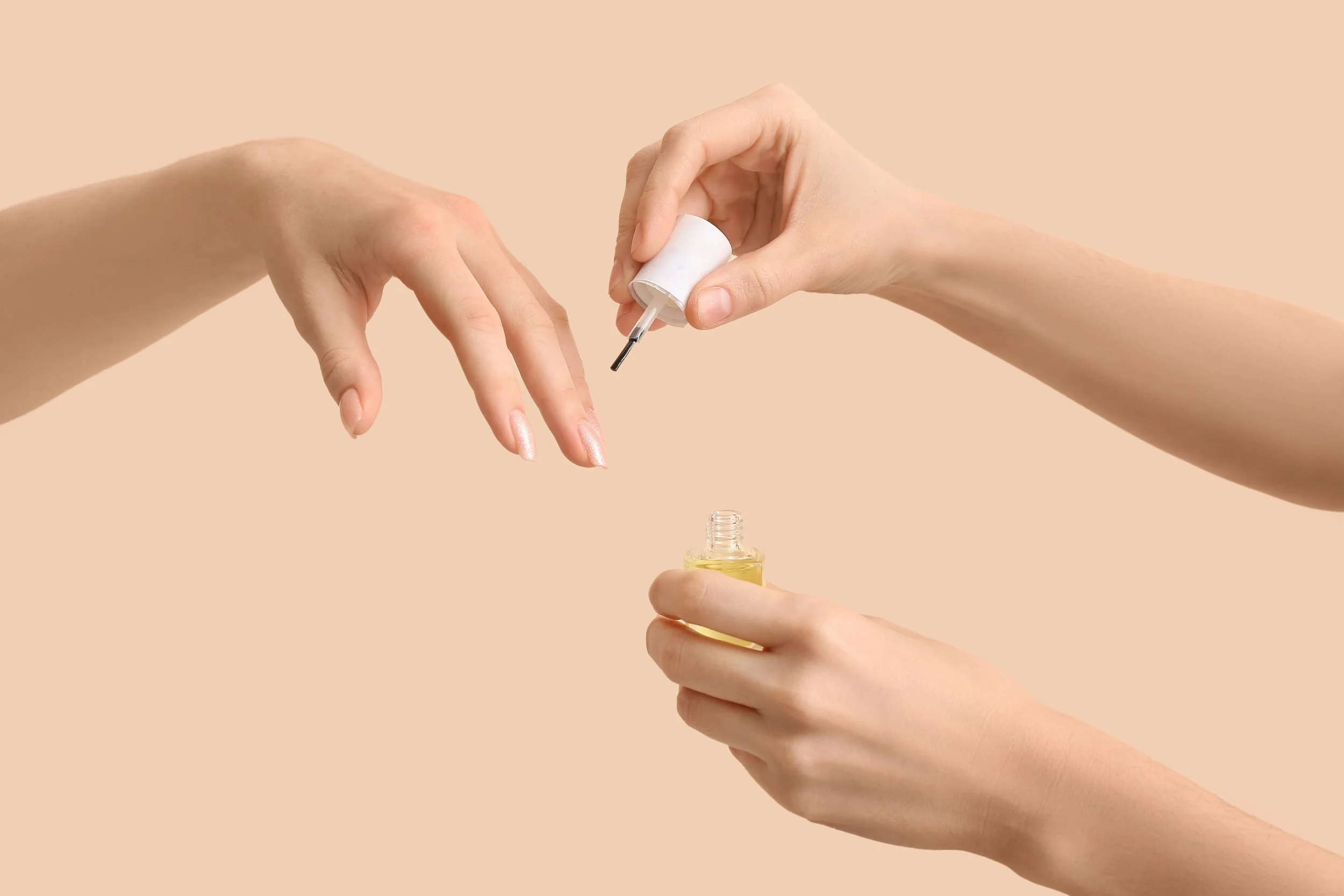 Two hands, one holding a small bottle of yellow oil and the other applying white nail polish from a small bottle with a brush.