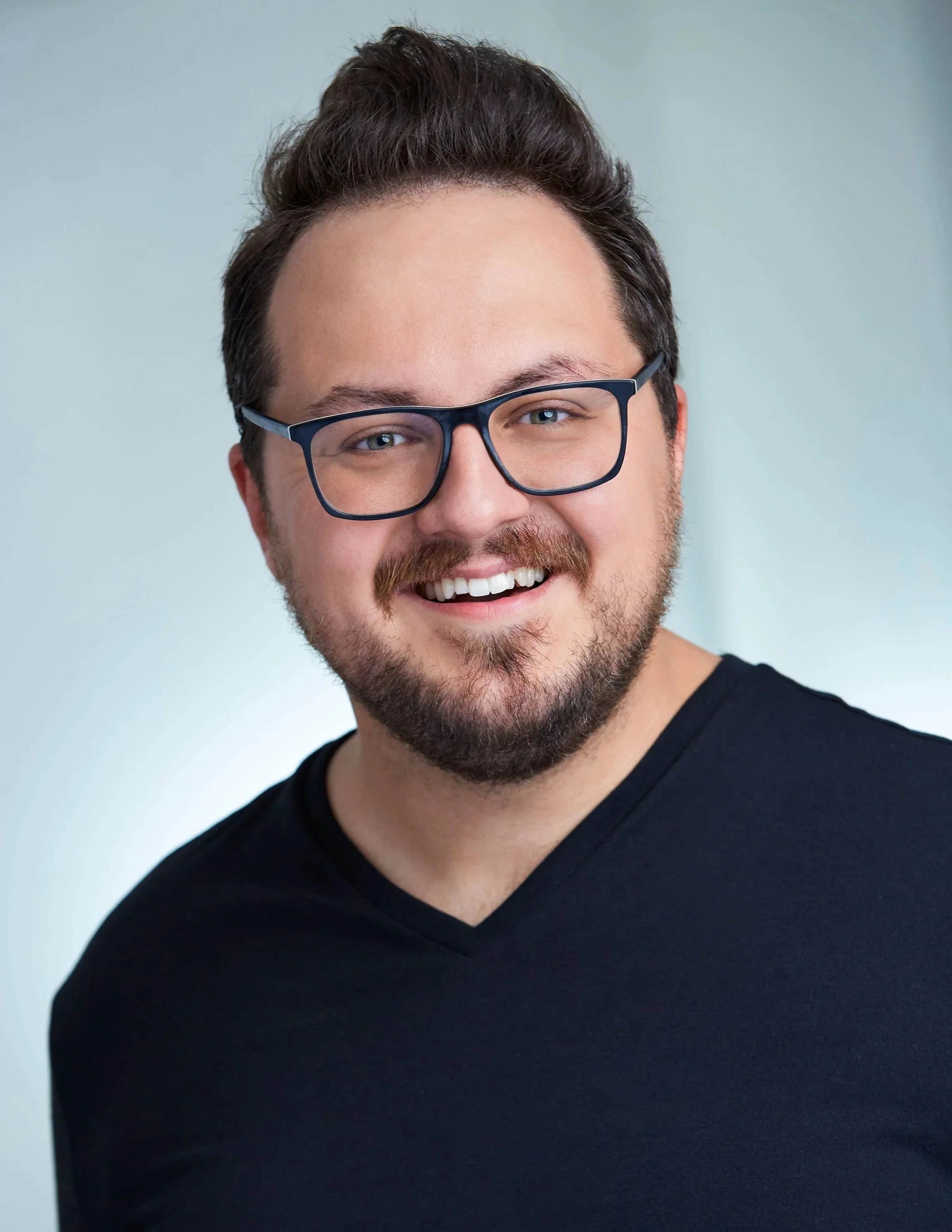 A smiling man with glasses, a beard, and dark hair, wearing a black shirt, posing against a light background.