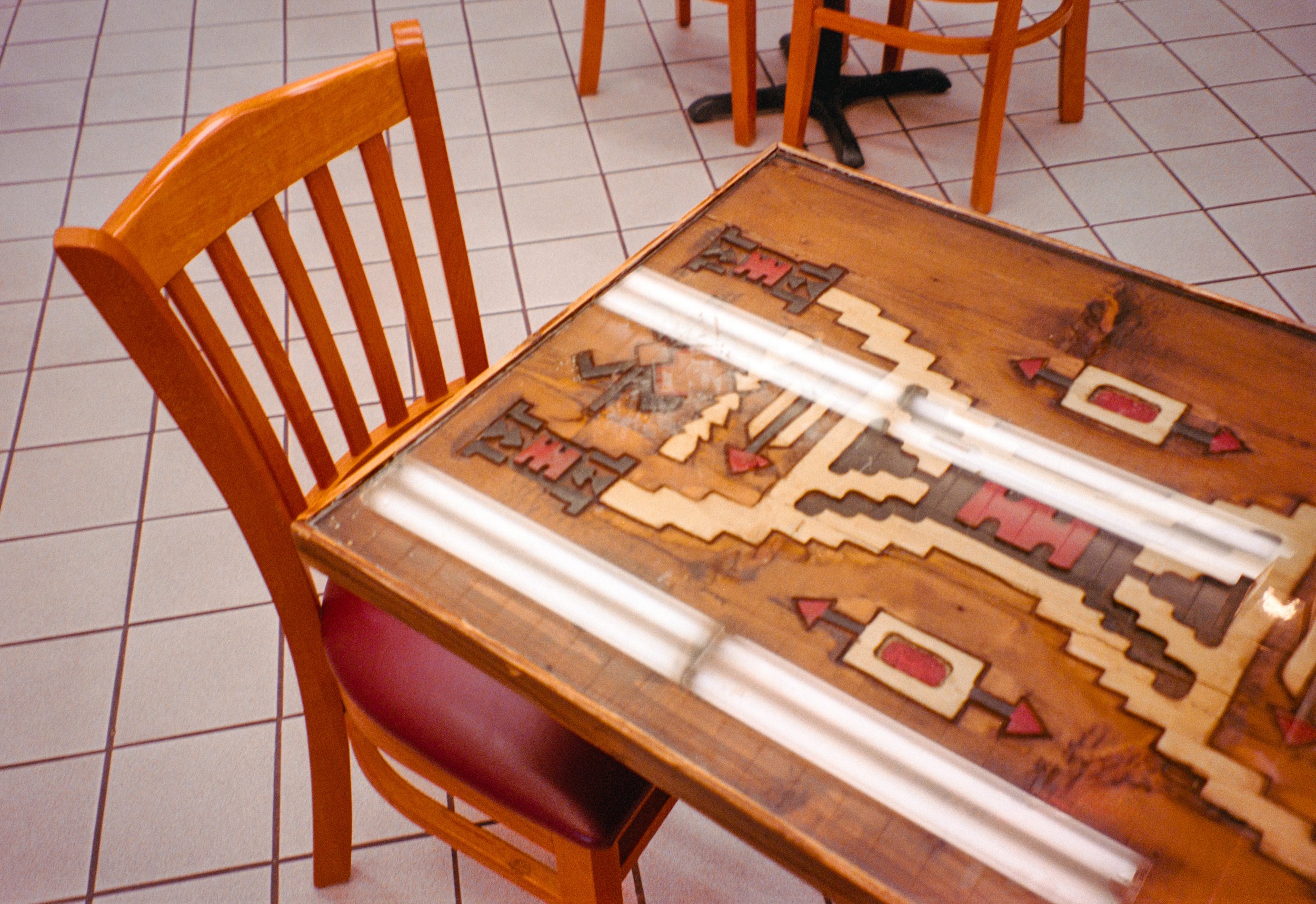 A wooden table with a glass top displaying a disassembled board game with wooden pieces, surrounded by wooden chairs, in a room with tiled floor.