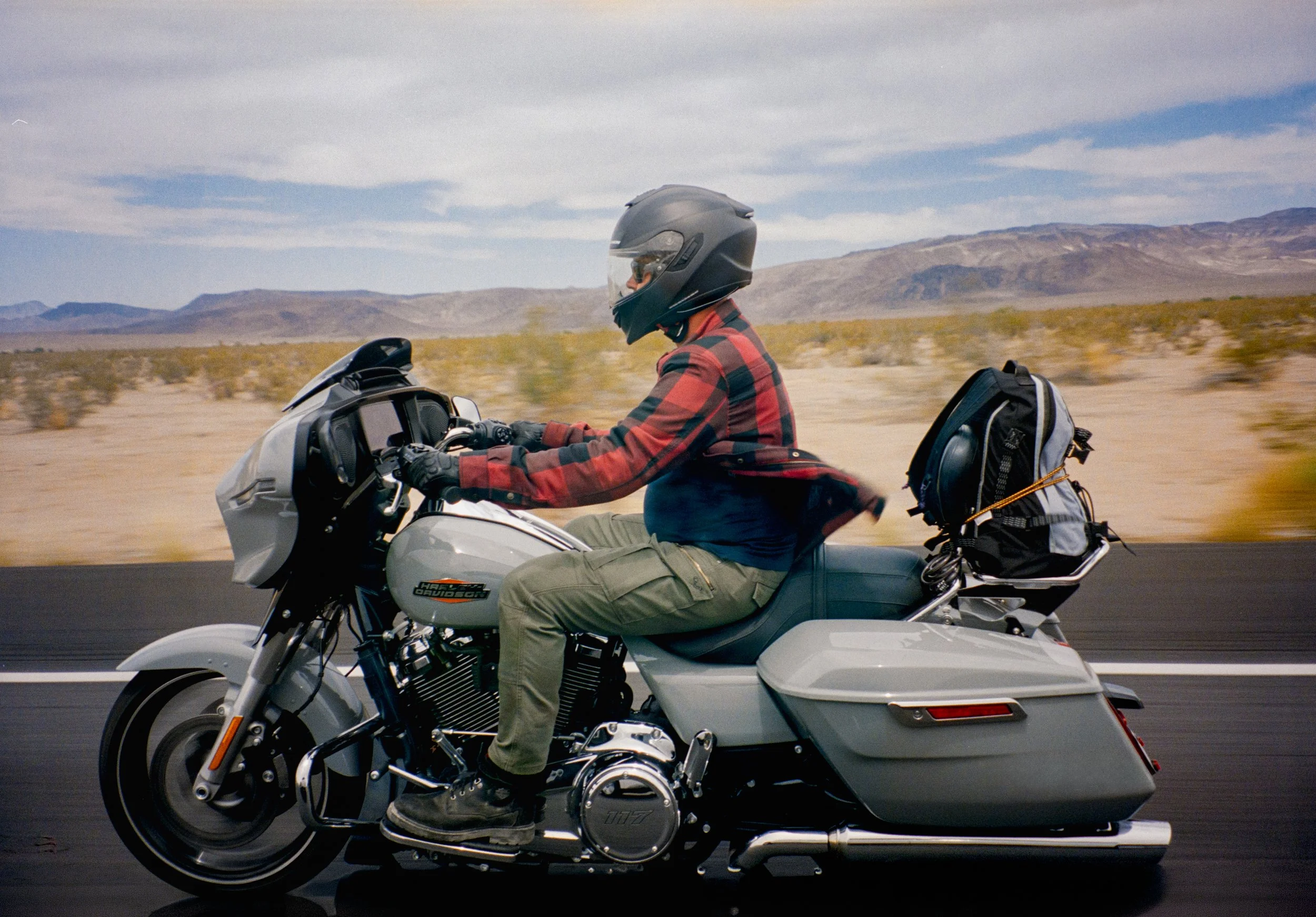A person wearing a helmet and plaid shirt riding a grey Harley Davidson motorcycle on a highway in a desert landscape with mountains in the background.