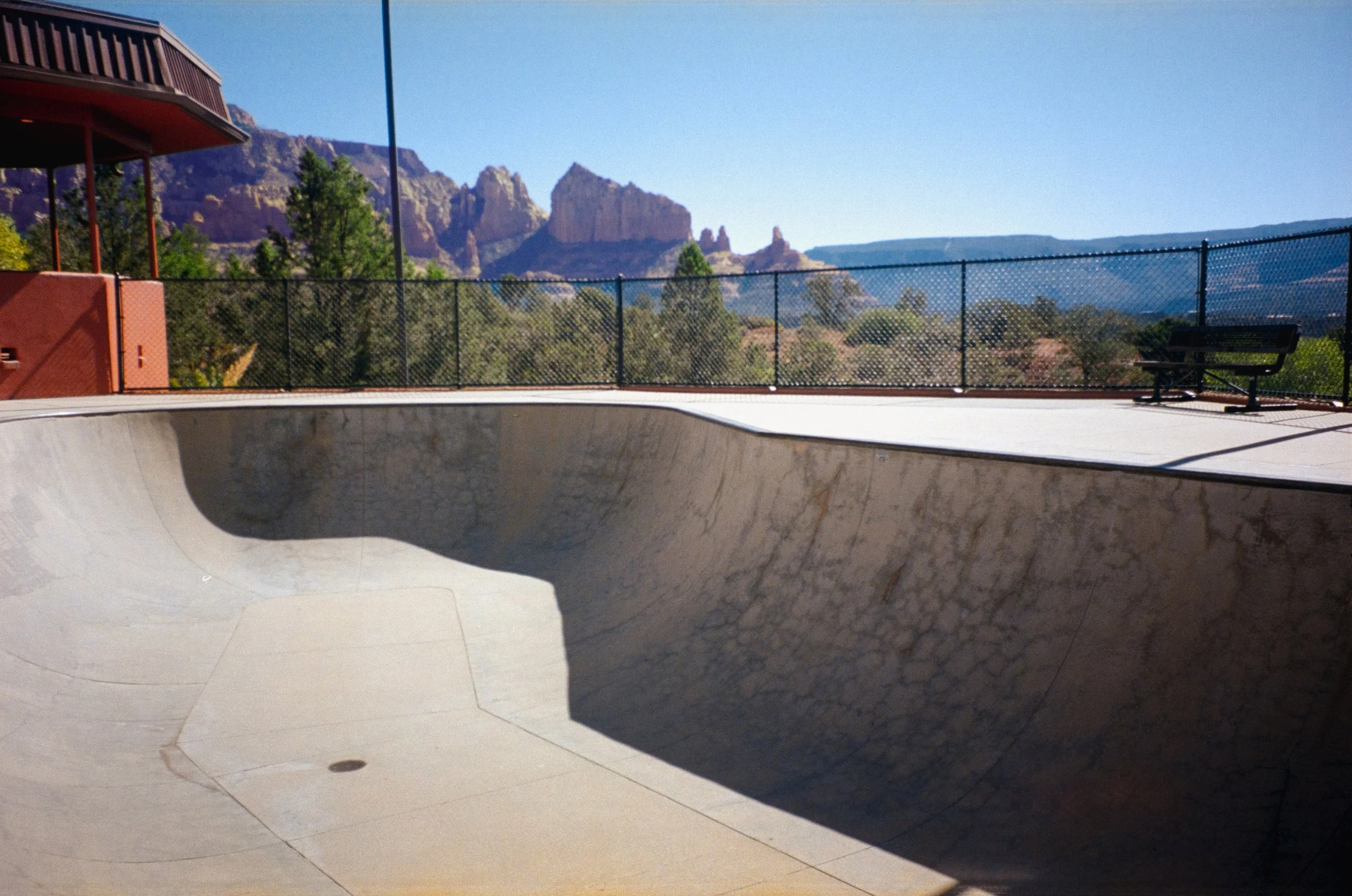 An empty outdoor concrete skate bowl with a chain-link fence surrounding it. In the background, there are trees and mountain formations under a clear blue sky.