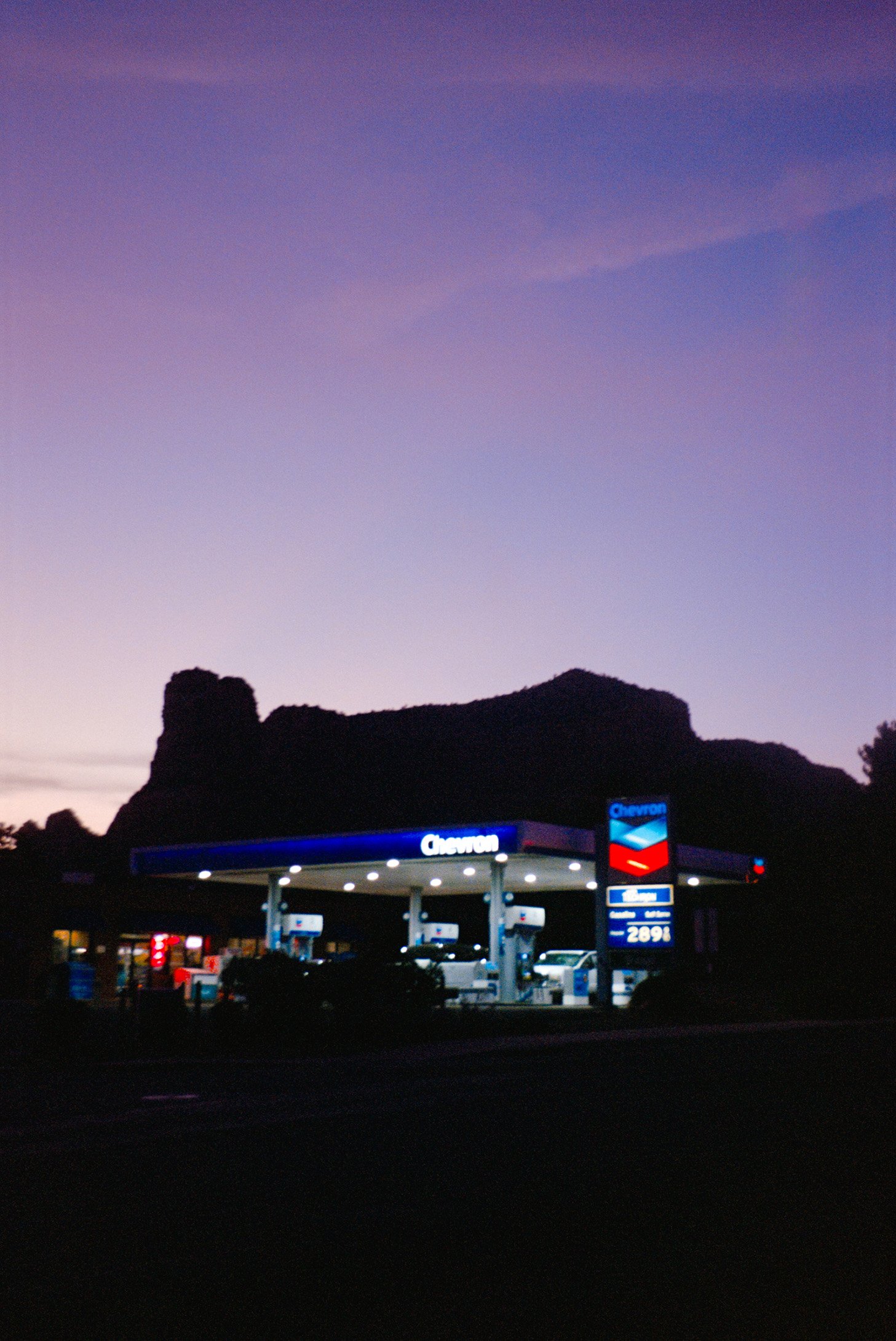 A Chevron gas station at dusk with a mountain in the background and a colorful purple and blue sky.