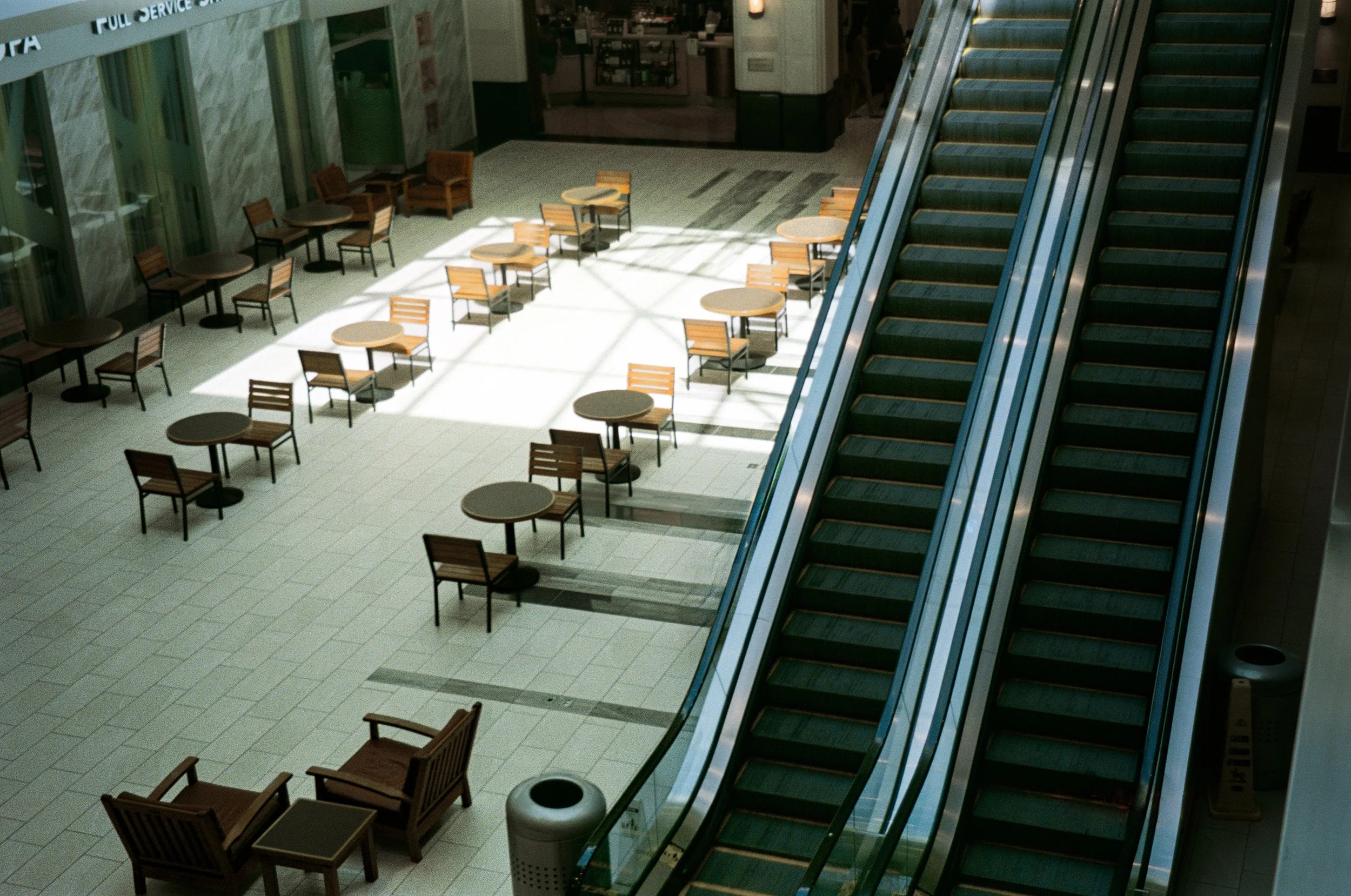 Empty indoor mall with escalators on the right and seating area with tables and chairs on the left, illuminated by sunlight coming through large windows.