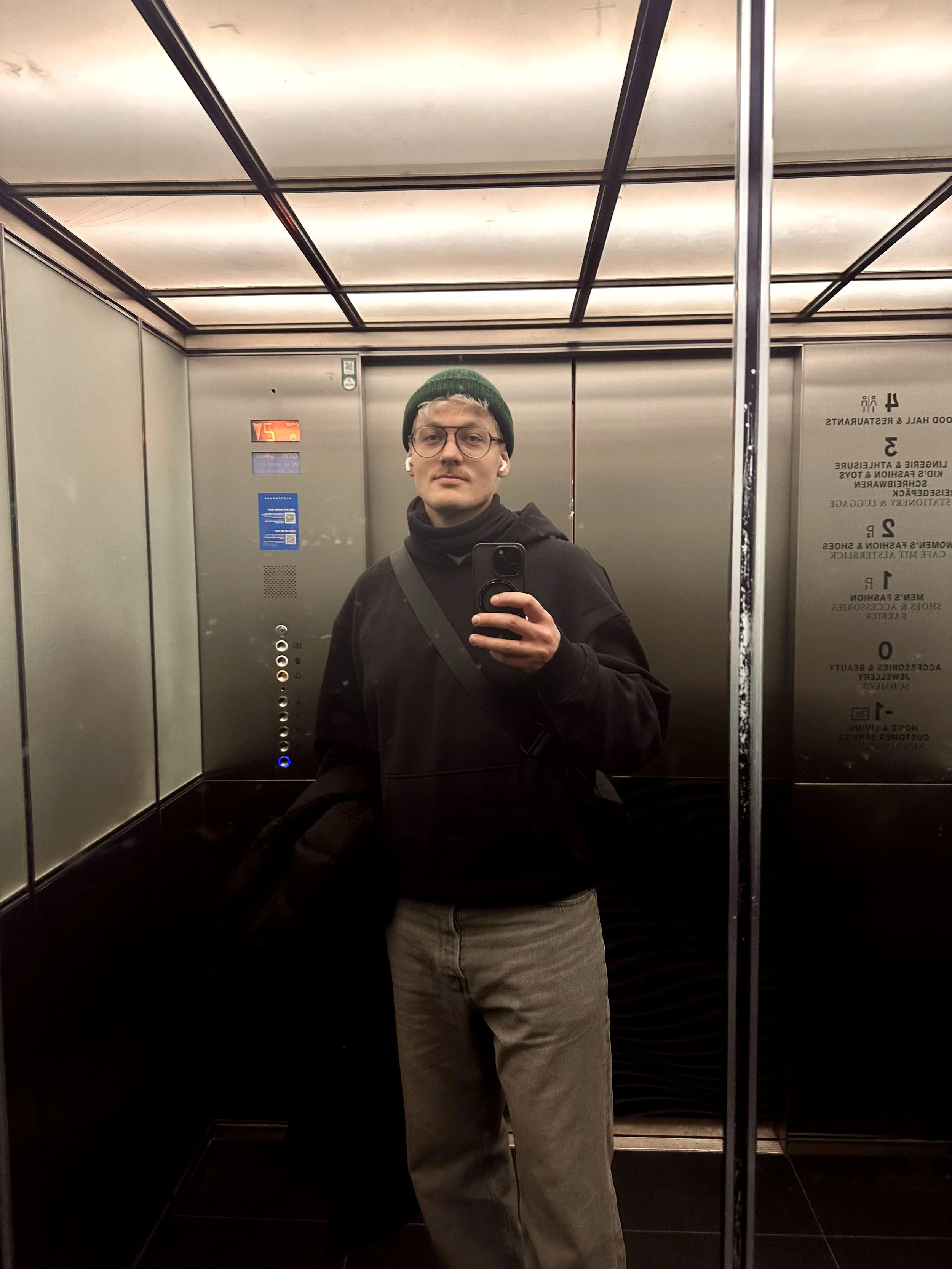 Junger Mann mit Brille, dunkelgrüner Mütze, schwarzen Jacke und beige Hose, nimmt Selfie in einem Aufzug mit Spiegel.