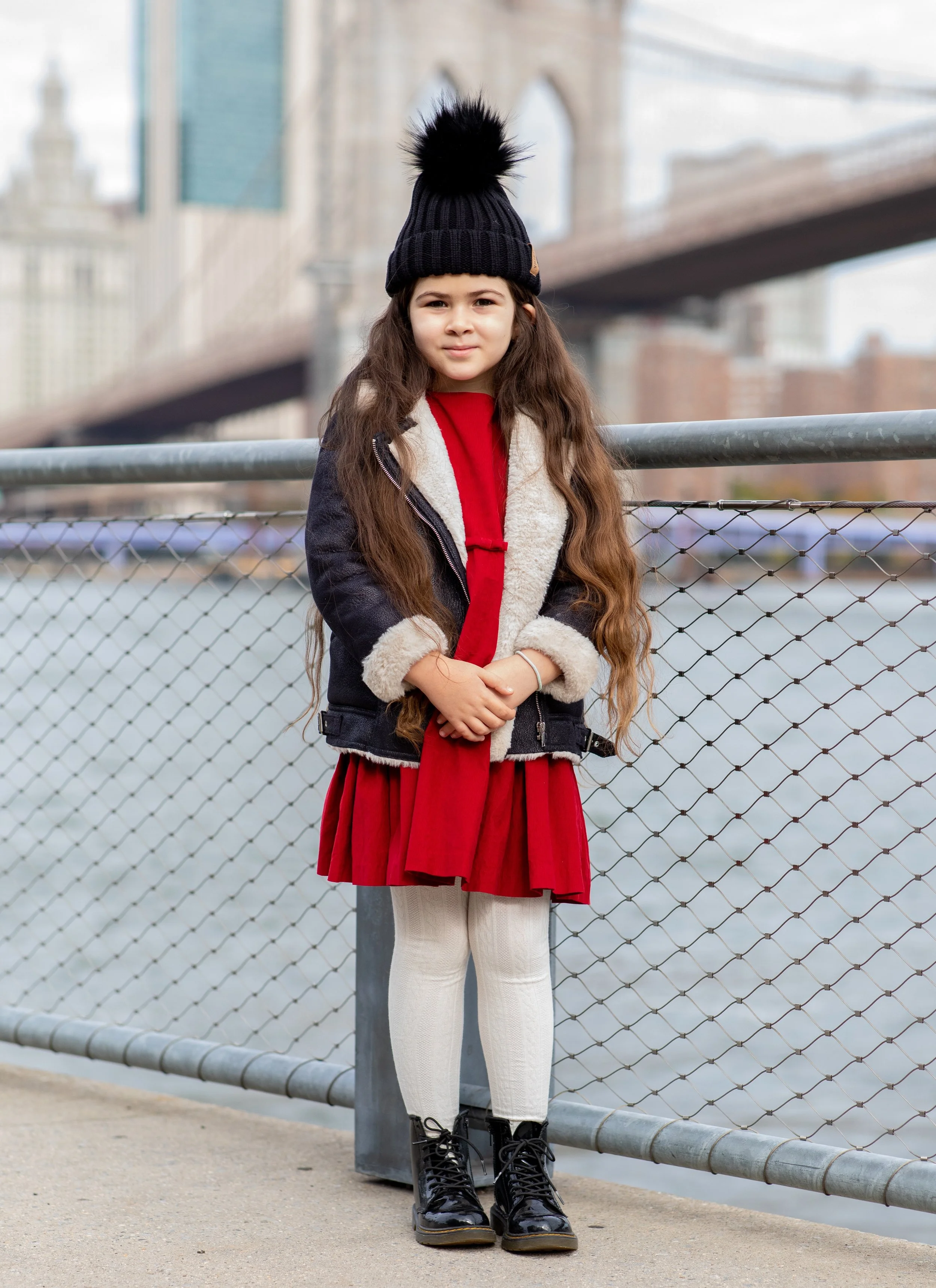 A young girl with long hair, wearing a black knit hat with a pom-pom, a denim jacket with fleece lining, a red dress, cream leggings, and black lace-up boots, standing near a metal fence with a cityscape and bridge in the background. MK Photo NYC