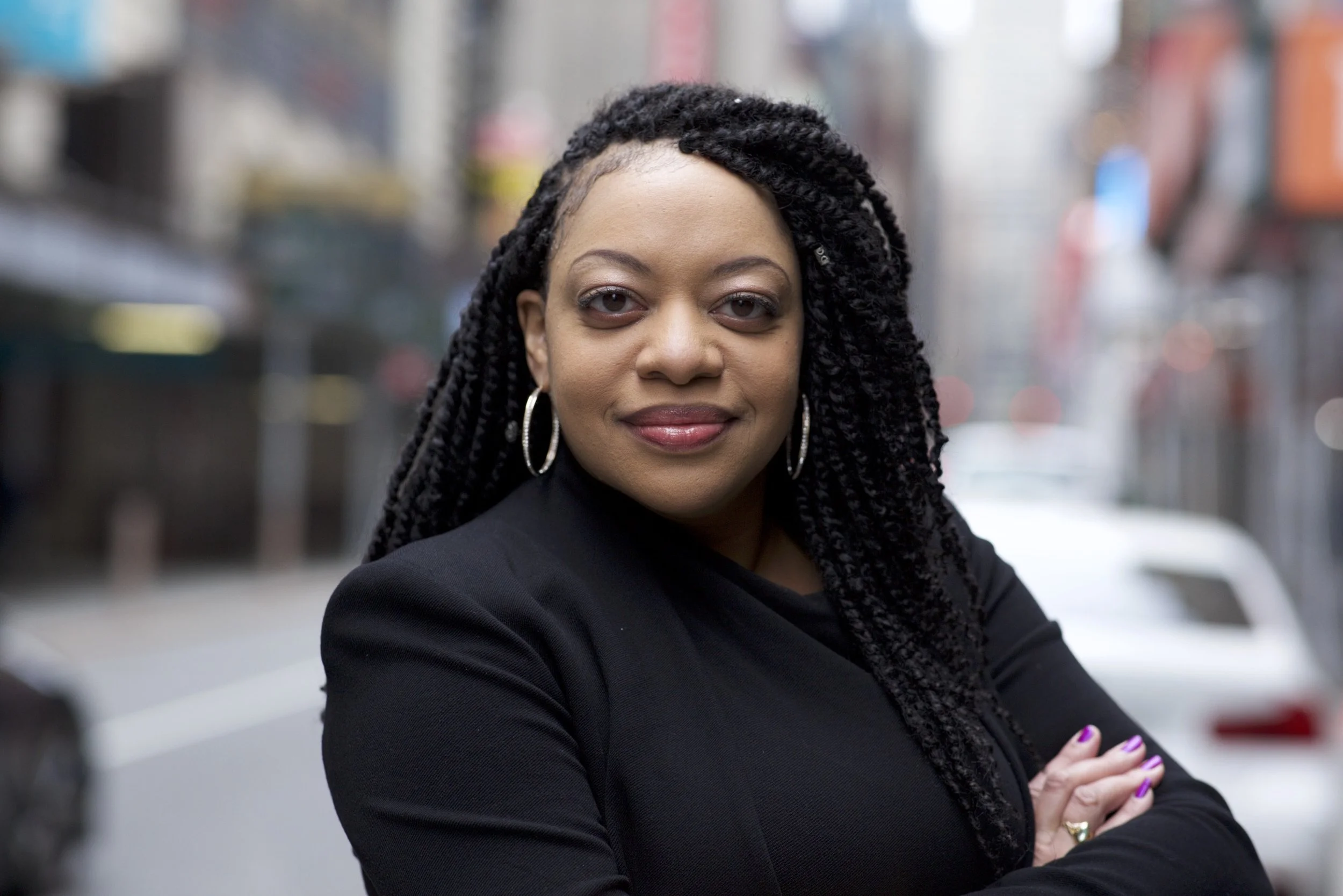 A woman with long, twisted black hair and hoop earrings, wearing a black jacket, standing outdoors in an urban setting with blurred buildings and cars in the background.