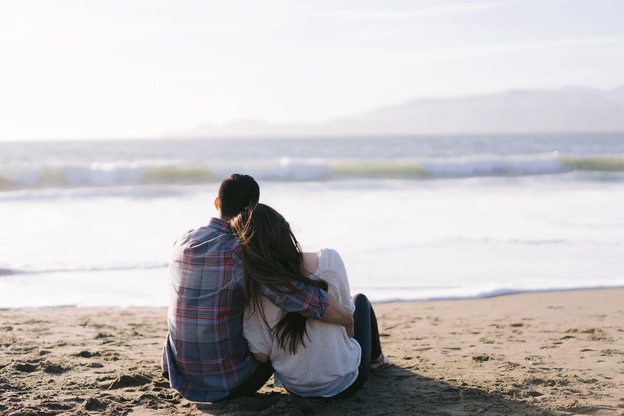 Engagement photo of couple in romantic cuddle watching the waves at sunset on Baker Beach in San Francisco.