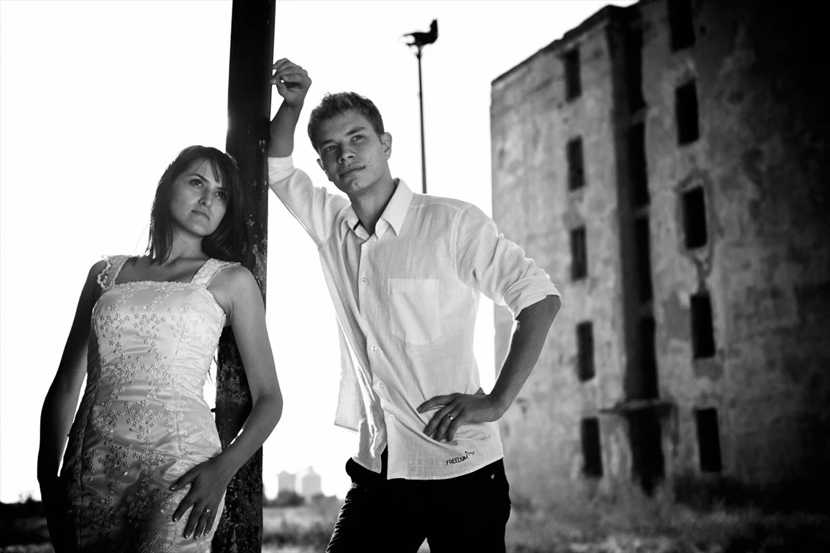 Black and white photo of a bride and groom standing next to a pole. The groom is in a white shirt, leaning against it, while the bride in a decorated wedding dress stands beside him. An industrial or abandoned building is visible in the background.