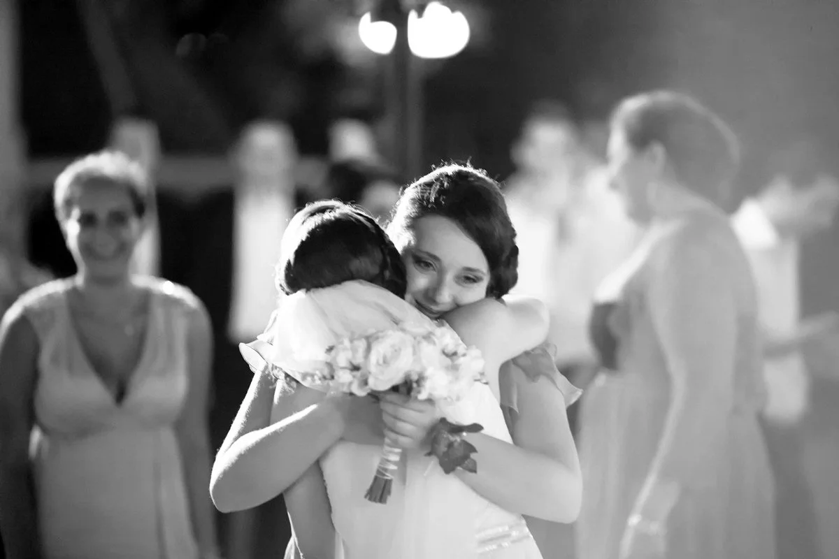 Bride and a friend hugging at a wedding, one holding a bouquet, with a happy crowd in the background.