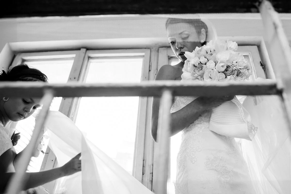 Bride holding bouquet of flowers next to a girl adjusting her veil, photographed from a low angle through a window frame.