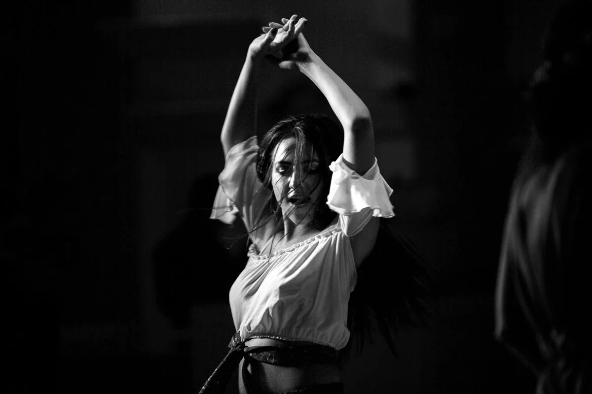 Dancer performing with arms raised, wearing a loose white blouse, in black and white.