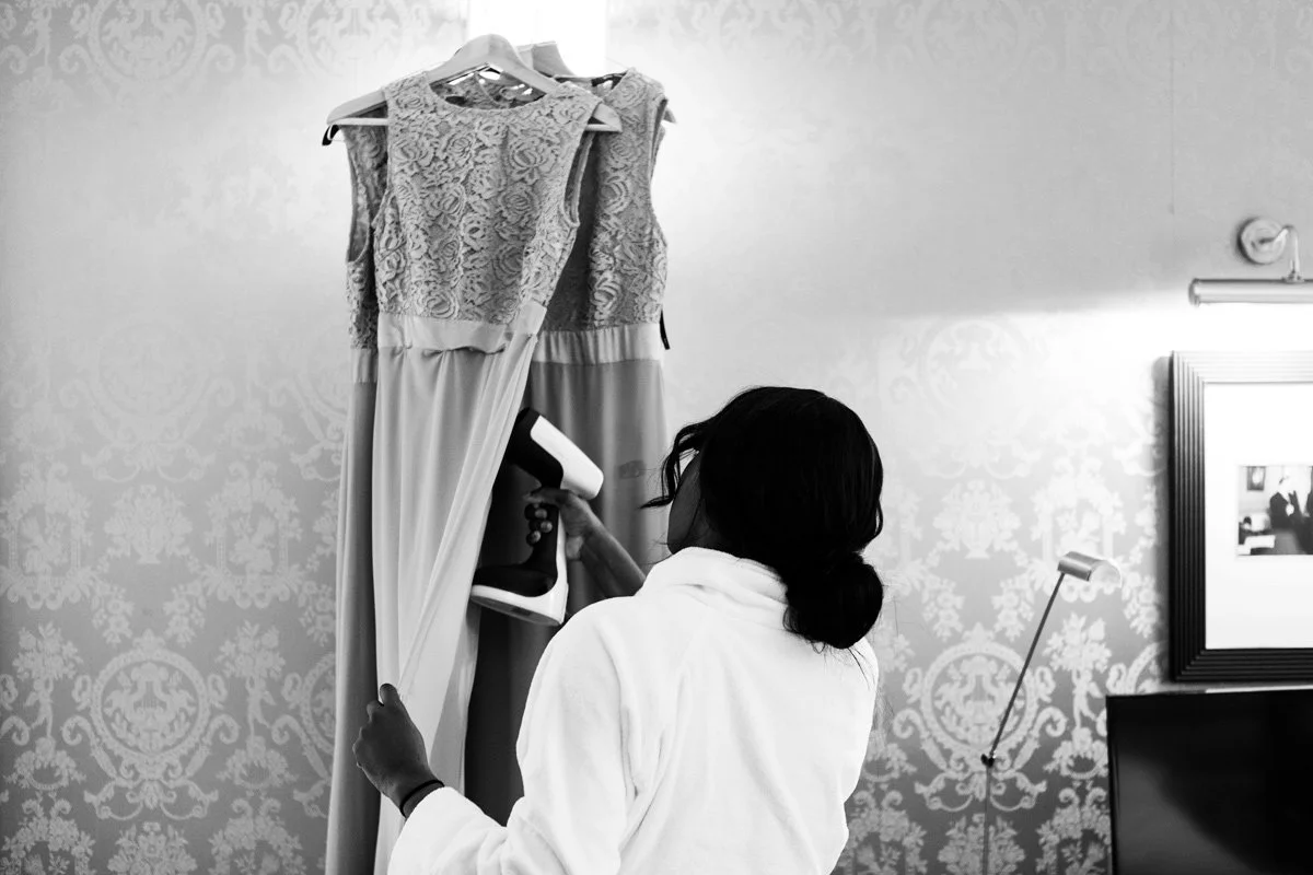 Bridesmaid steaming a lace dress with a handheld garment steamer in a room with patterned wallpaper.