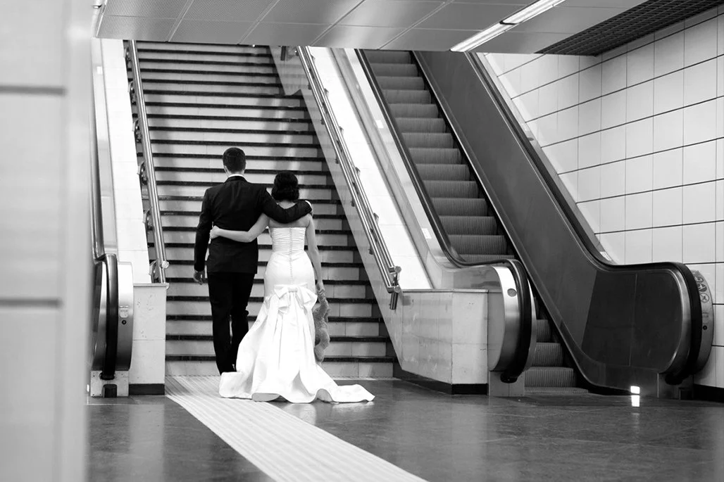A couple in formal attire walking arm in arm up stairs next to stationary escalators.