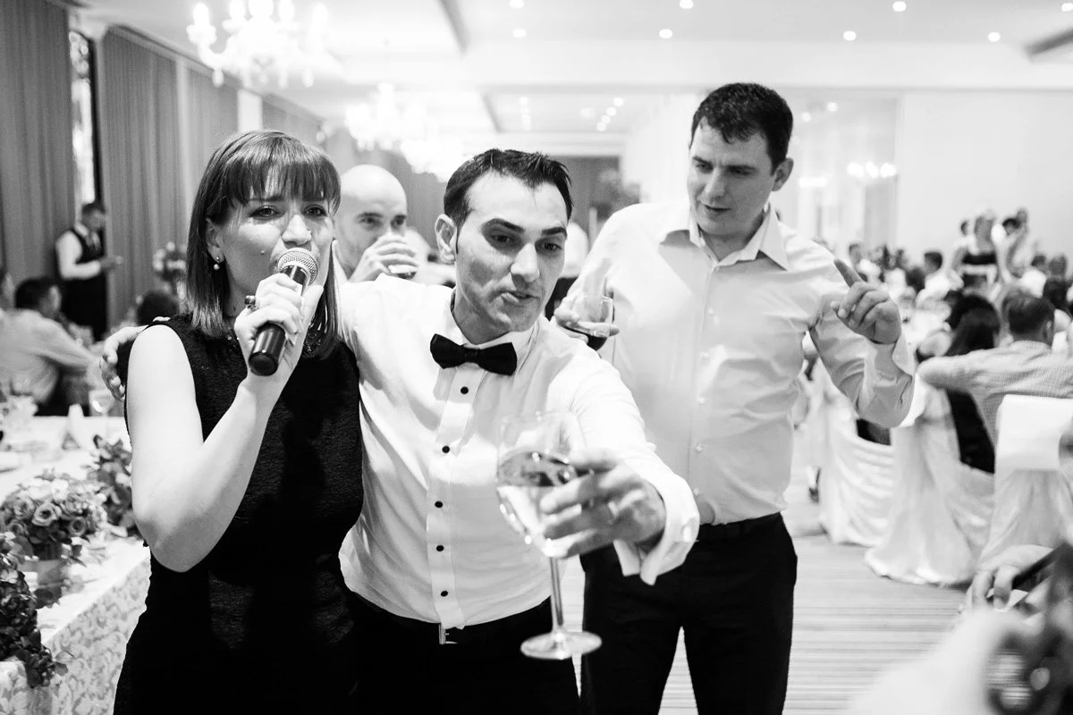 People celebrating at a wedding party, one holding a microphone and others with drinks, in a banquet hall.