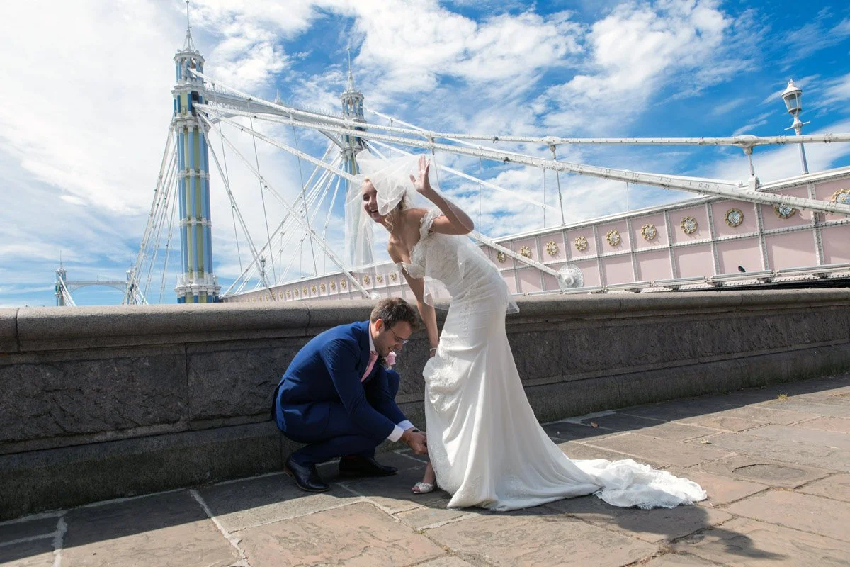 Bride and groom on a bridge, groom adjusting the bride's shoe, with a decorative bridge structure in the background and blue sky.