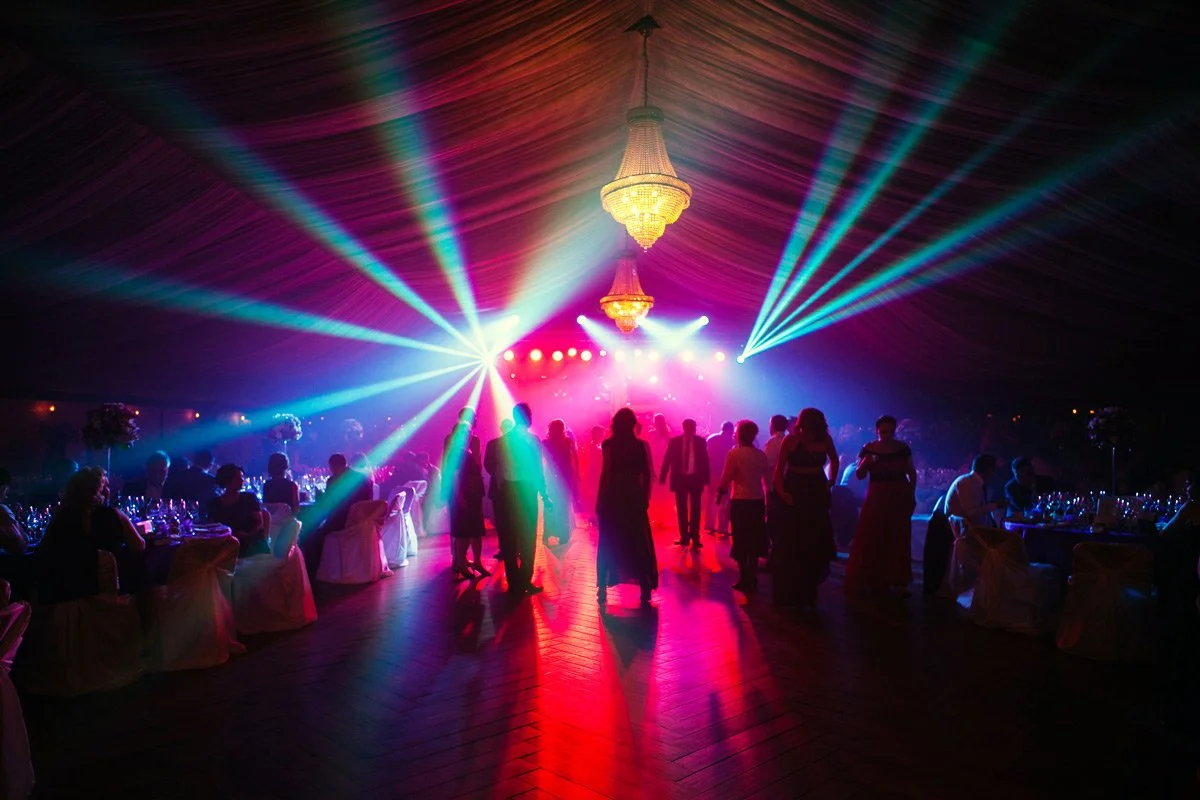 People dancing and socialising at a wedding party with colourful lighting and a chandelier overhead.