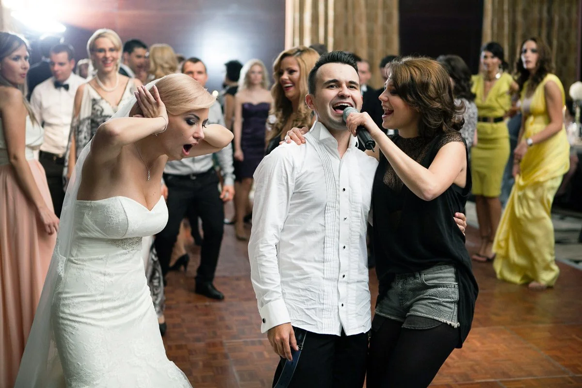A bride in a white gown covering her ears while two people sing into a microphone at a lively wedding reception.