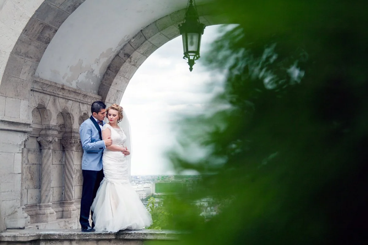 Bride and groom embracing under an arched stone structure, with greenery in the foreground.