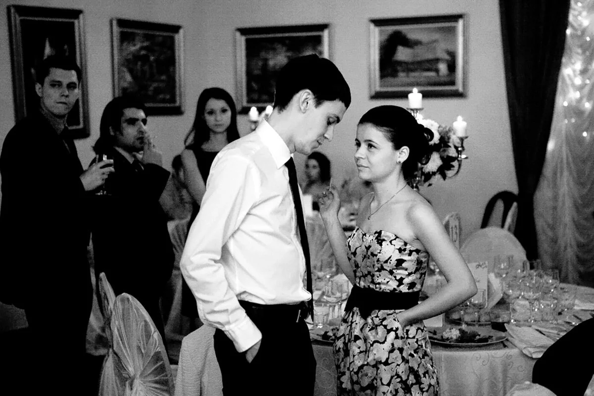 Group of people in formal attire at a wedding event, with a man and woman conversing closely, surrounded by tables with tableware and decorations, and others observing in the background.