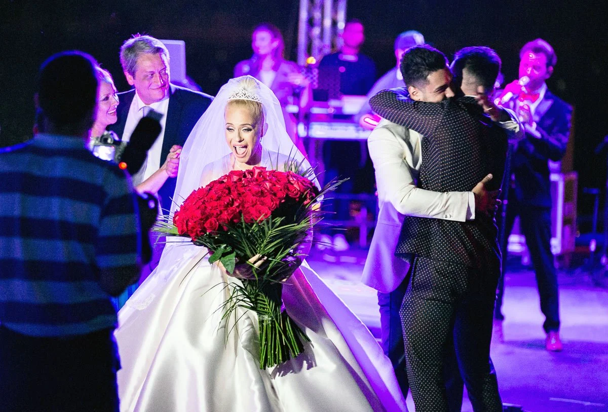 Bride holding a large bouquet of red roses, surrounded by people at a wedding celebration, with a band in the background and a person photographing the scene.