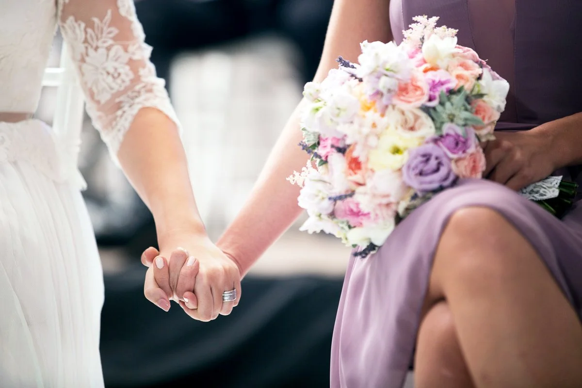 Bridesmaid and bride holding hands, one in a white lace dress and the other in a lavender dress holding a colorful bouquet.