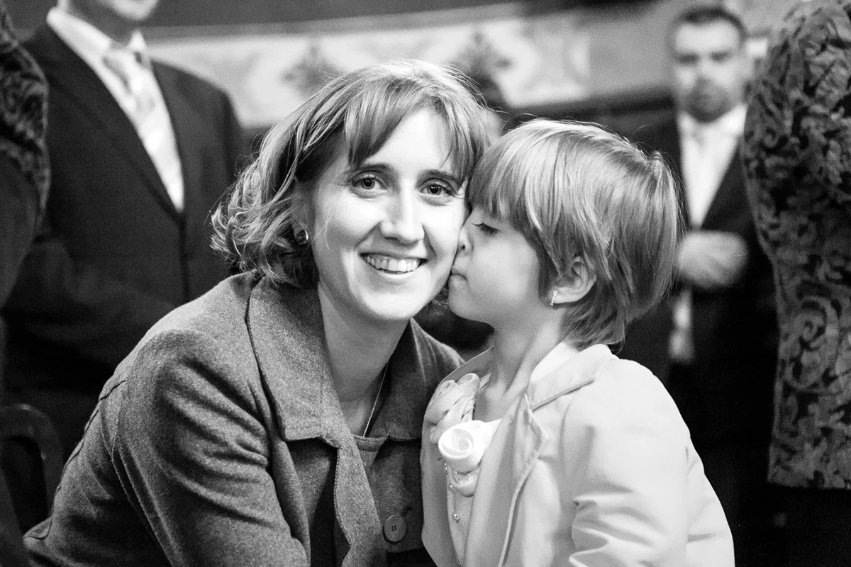 A woman smiling as a child kisses her cheek, in black and white.