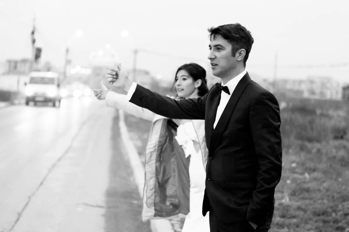 Black and white photo of a bride and groom in formal attire hitchhiking on a road.