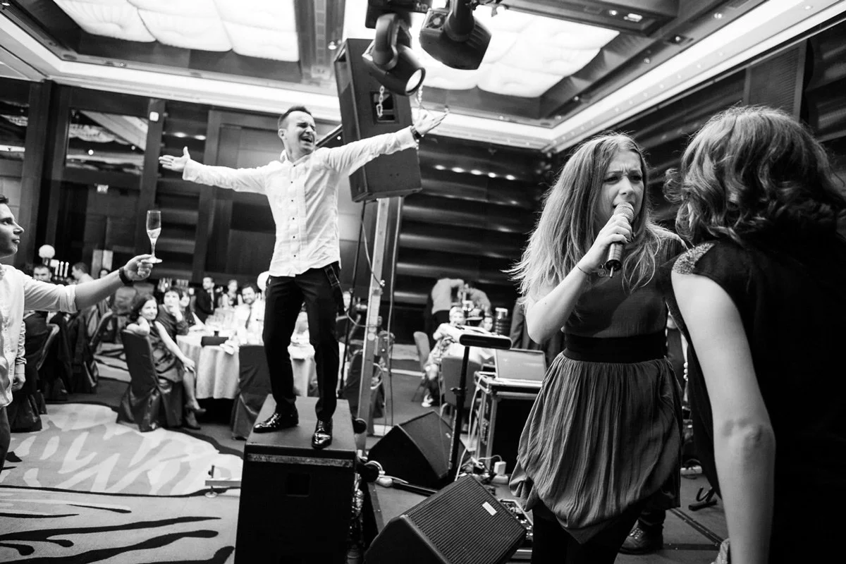 A lively party scene with people singing and dancing. A woman is singing into a microphone. The scene is energetic, with guests seated at tables in the background, and one person toasting with a glass.