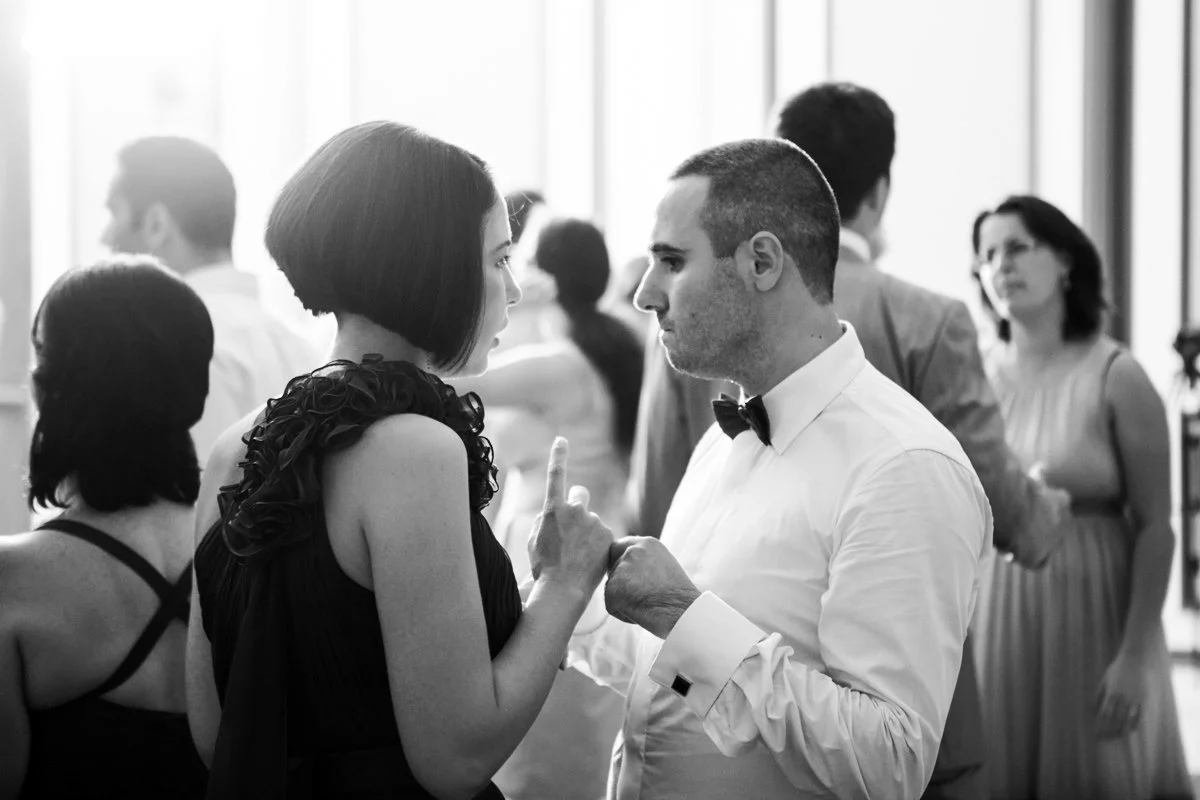 Black and white photo of a man and woman in formal attire conversing at a wedding, with people in the background.