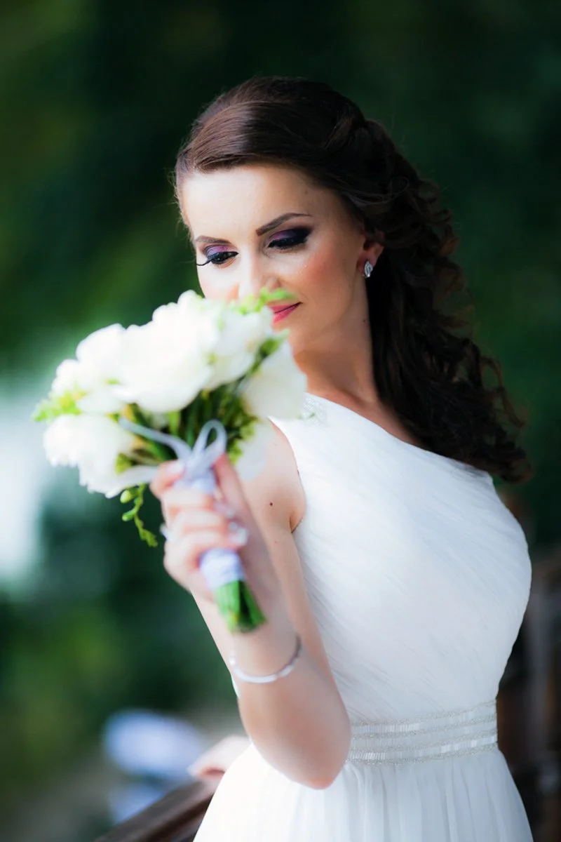 Bride holding a white bouquet, smiling gently, in a white wedding dress with greenery background.