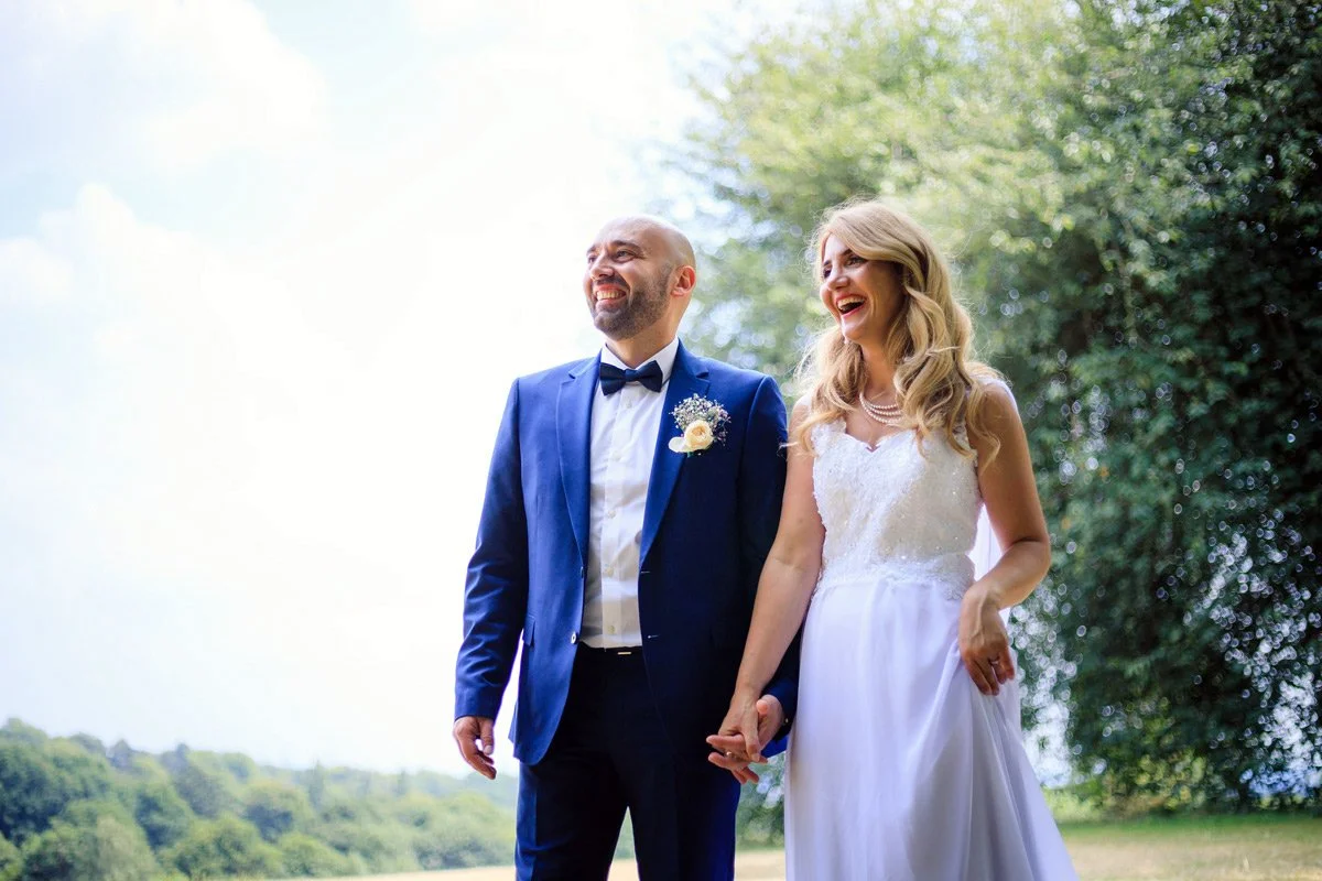 A bride and groom holding hands and smiling outdoors, with trees and sky in the background.