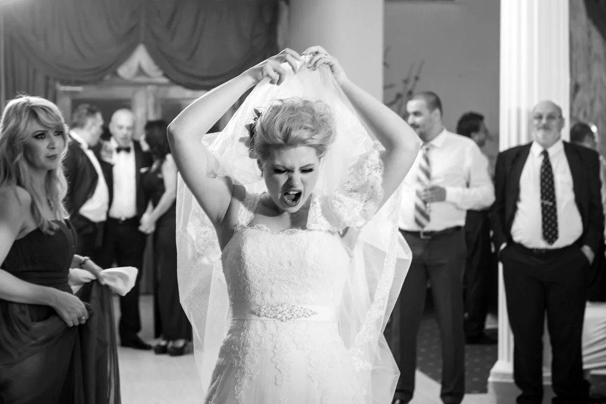 Bride lifting veil and making an expressive face at a wedding reception, surrounded by guests.