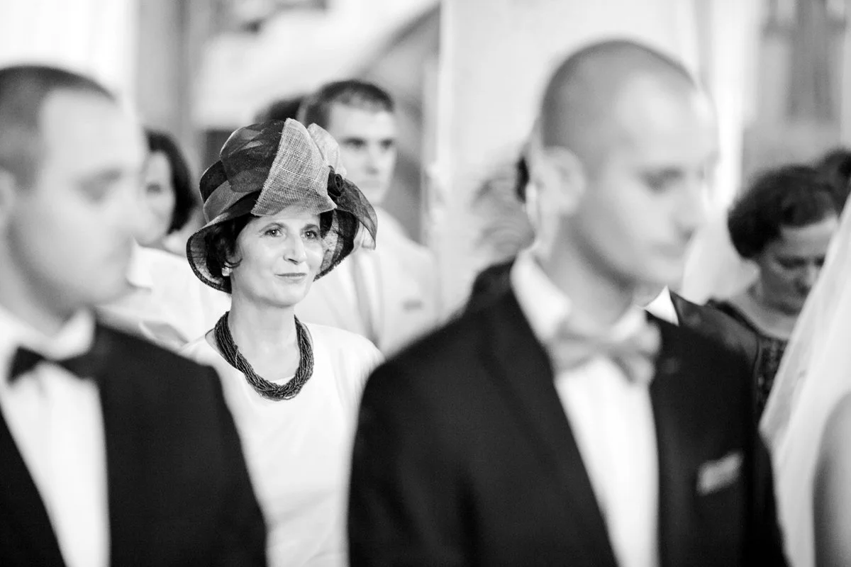 Black and white image of the mother of the groom in a hat at a wedding ceremony. She is surrounded by other attendees, with a focus on her expression.