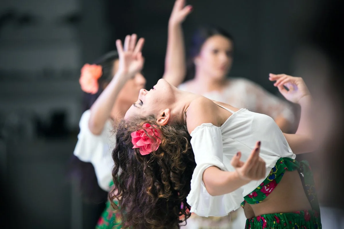 Dancers performing with one in focus wearing white floral attire, highlighting a graceful pose with arms raised and a pink flower in her curly hair.