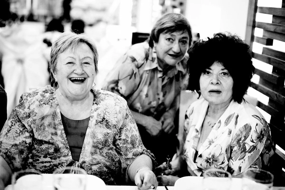 Three elderly women smiling and posing for a black-and-white photo in a dining setting.