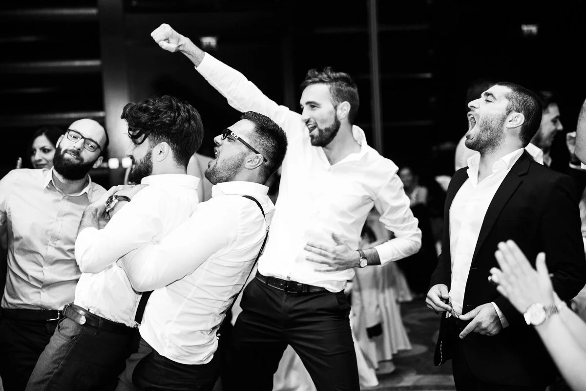 A group of five men in formal attire celebrating, with some smiling and one raising a fist, at a wedding party.