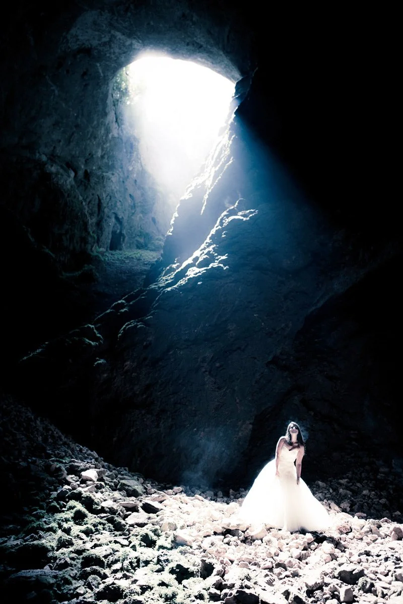 Bride standing inside a cave with sunlight streaming in from the opening above.