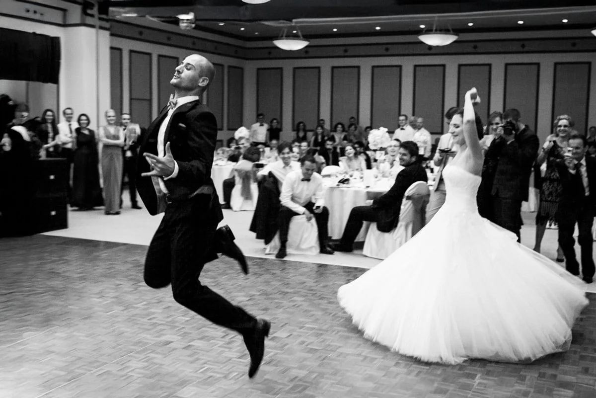 A bride and groom dancing exuberantly at a wedding reception, surrounded by guests seated and standing, in a banquet hall setting.