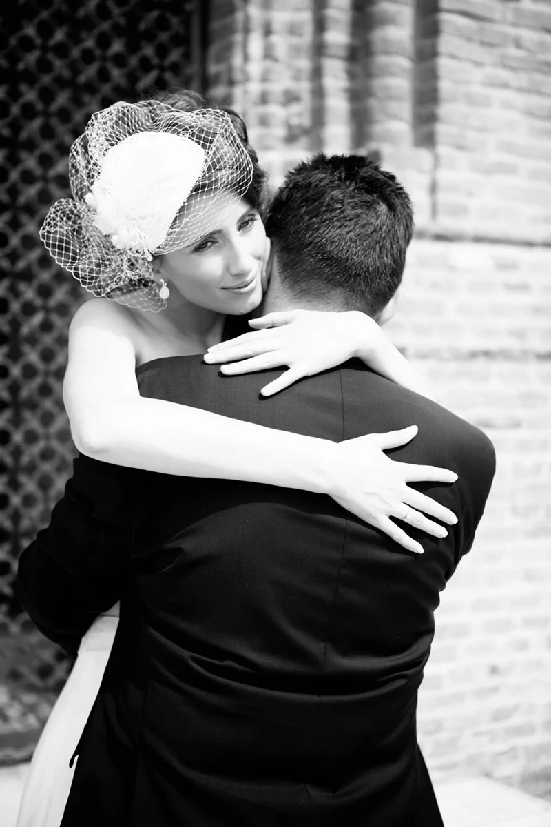 A bride embracing the groom in a black and white photo, with the bride wearing a veil and the groom in a suit, in front of a brick wall.