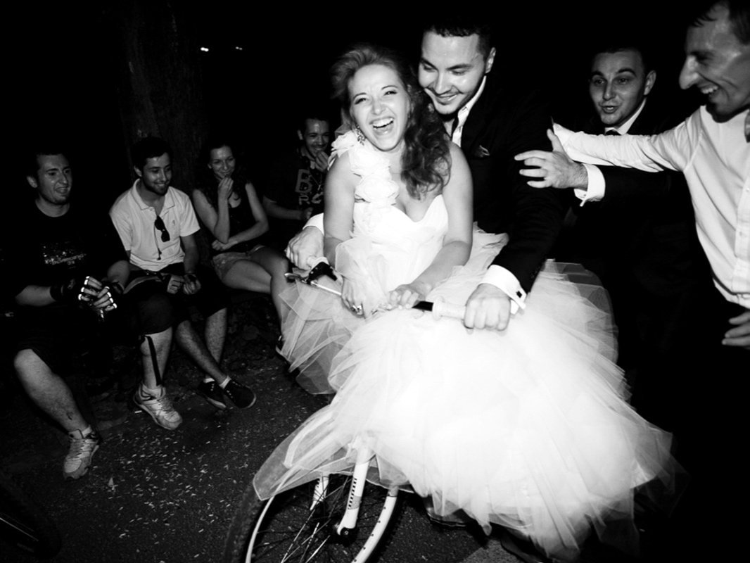 Bride and groom in formal attire laughing while riding a bicycle surrounded by friends in a playful setting.