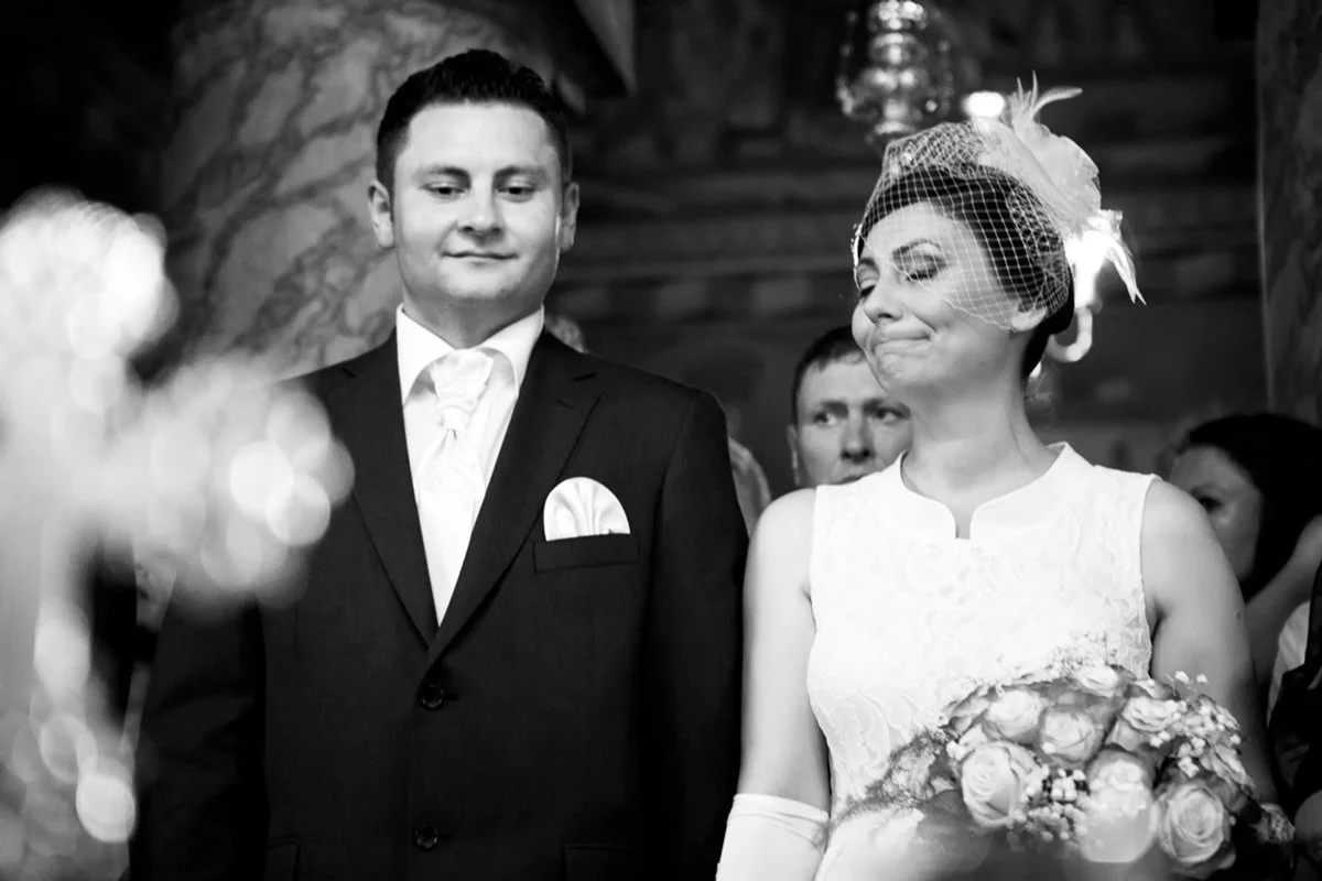 A black and white photo of a wedding ceremony with a groom in a suit and a bride wearing a lace dress and a fascinator, holding a bouquet.